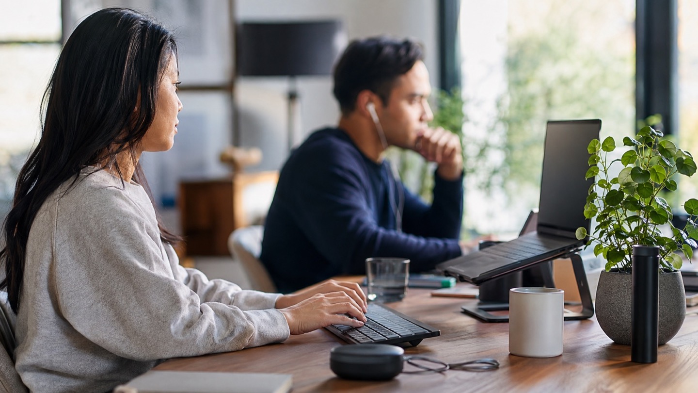 Person typing on a quiet keyboard at a shared workspace with another person nearby